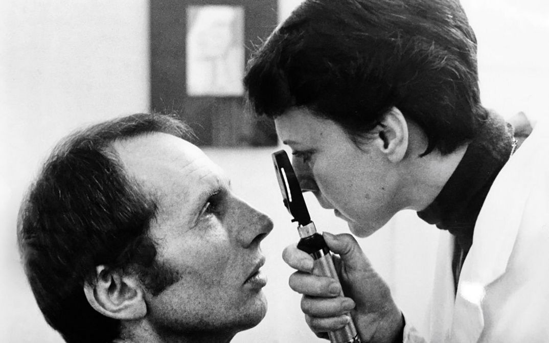 Black and white image of a middle-aged man and a female doctor, both are in profile to the camera. The doctor is looking slightly down at the sitting man, studying his eye with a magnifying lens.