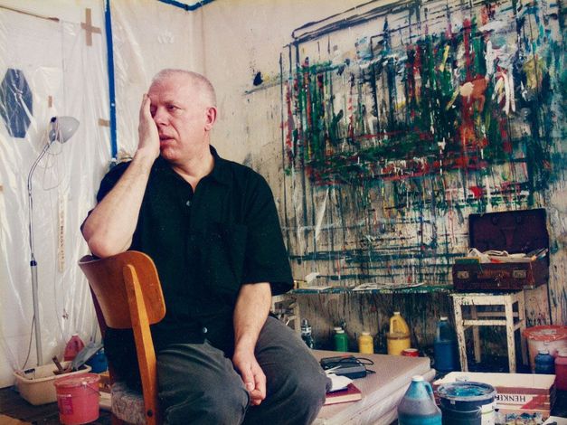 Künstler sitzt im Atelier vor bemalter Wand, Hand im Gesicht.