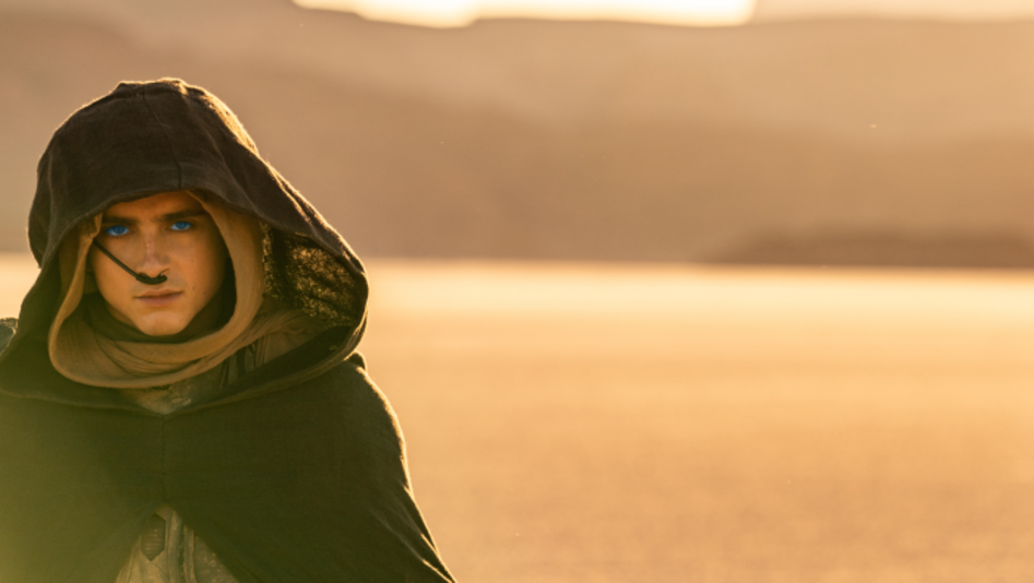Filmstill from DUNE 2: A young man in the desert. He is wearing a cloak with a hood, has very blue eyes and a tube on his nose.