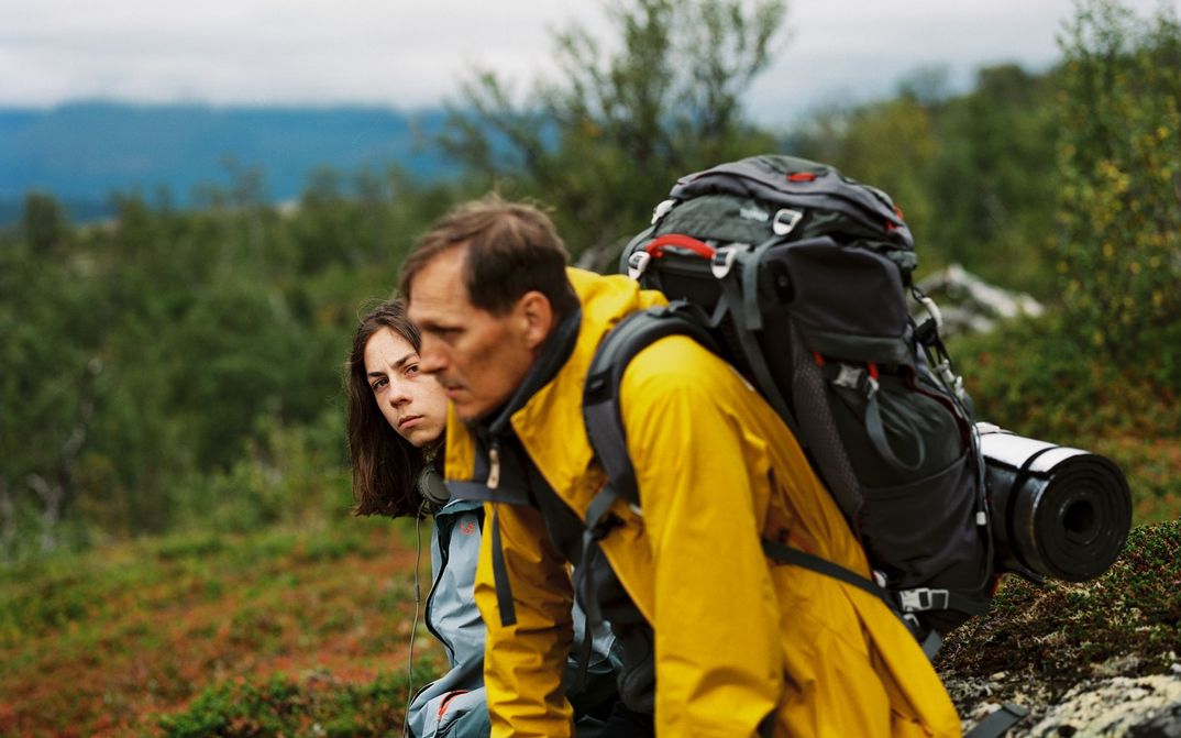 Filmstill from HELLE NÄCHTE: A man with a hiking rucksack and sleeping mat and another younger person are sitting on a rock in the mountains. Trees and rocks can be seen.