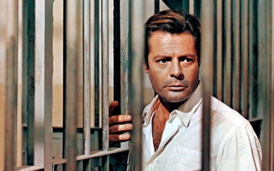 Film still from LO STRANIERO: Marcello Mastroianni is seen between the bars of a prison.