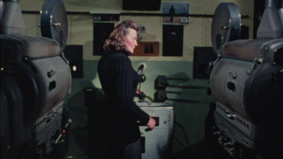 Film still aus RAIN OR SHINE: A woman stands between two 35mm projectors in a projection booth.