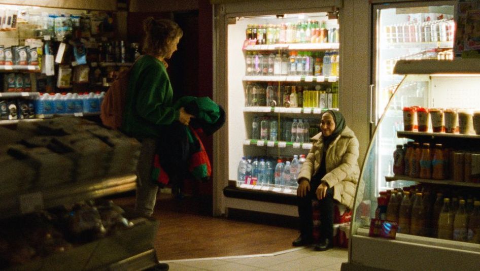 Film still from GHOST TROPIC: Two women can be seen smiling at each other in a dimly lit supermarket.