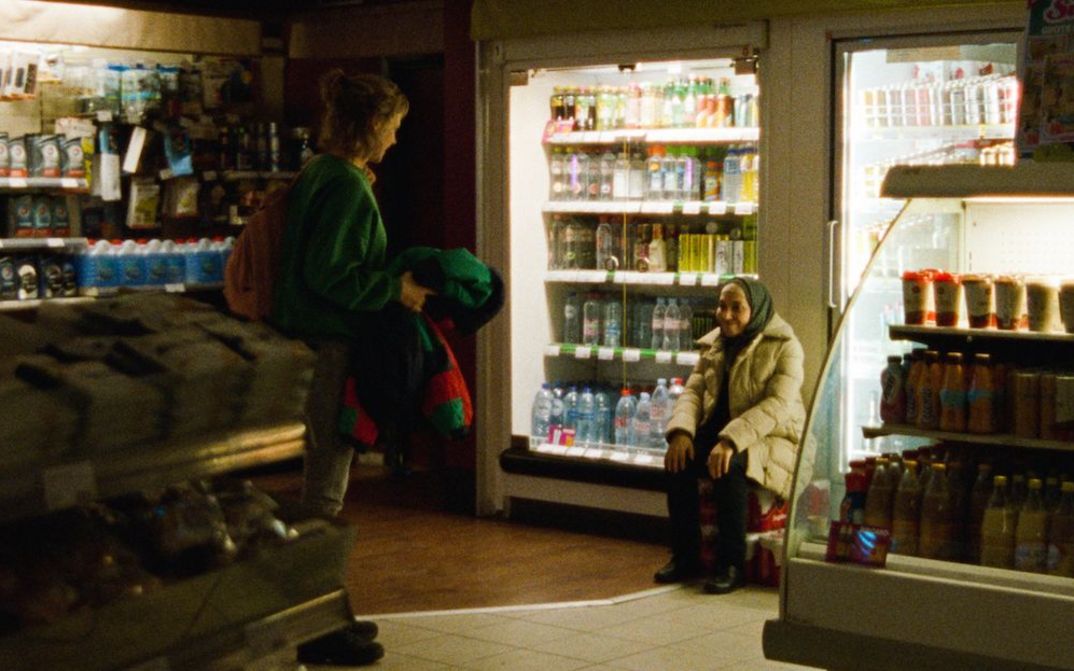 Film still from GHOST TROPIC: Two women can be seen smiling at each other in a dimly lit supermarket.