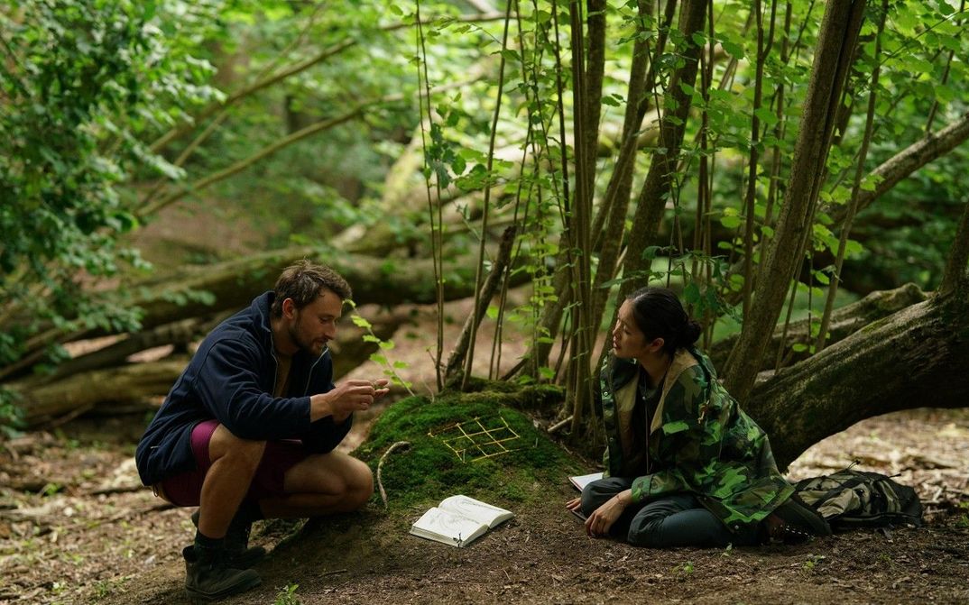 Film still from HERE: Two people sit on the forest floor and look at the moss between them.