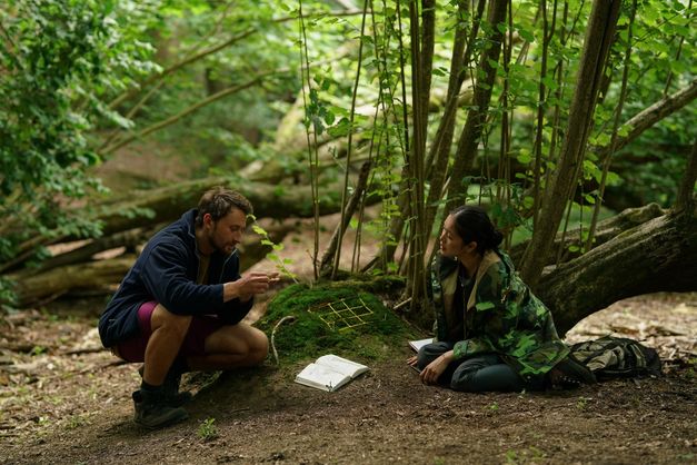 Film still from HERE: Two people sit on the forest floor and look at the moss between them.