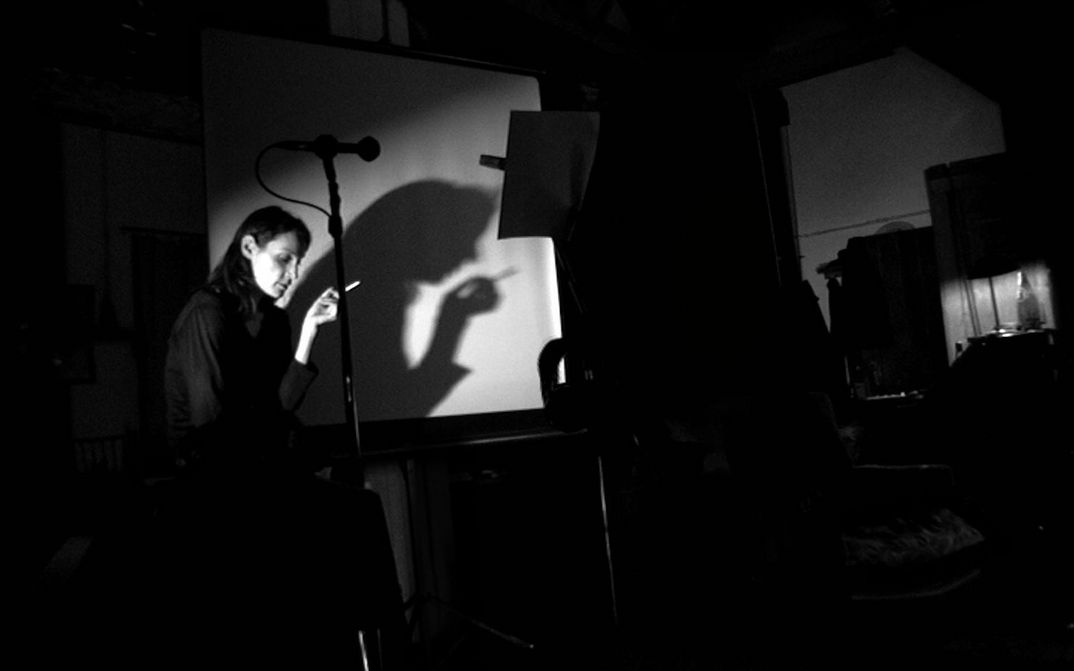 Film still from NE CHANGE RIEN: In a dark room, a woman stands in front of a screen with a cigarette in her hand. In front of her are a microphone and a music stand.