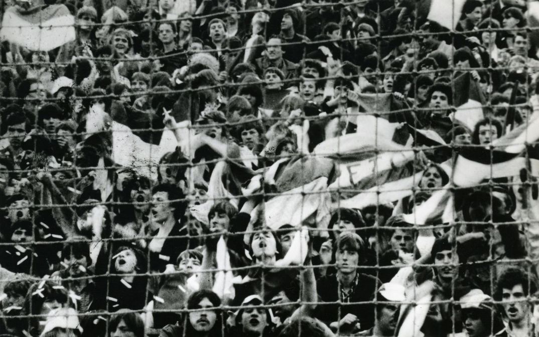 Film still from ARÉNA. In black and white you see cheering spectators of a sports event, they are behind a fence.