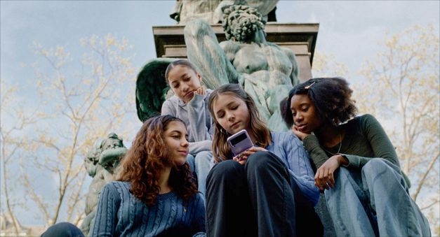 Filmstill aus WENN DU ANGST HAST NIMMST DU DEIN HERZ IN DEN MUND UND LÄCHELST von Marie Luise Lehner.  Vier junge Frauen sitzen vor einer Statue, alles schauen auf das Handy, das die mitlere von ihnen  in der Hand hält. 