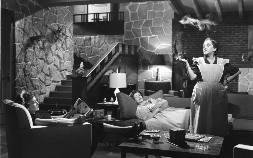 Film still from EL CASO DE LA MUJER ASESINADITA: Two women are sitting and lying on the couch in a living room. A maid stands in the middle of the room smoking.