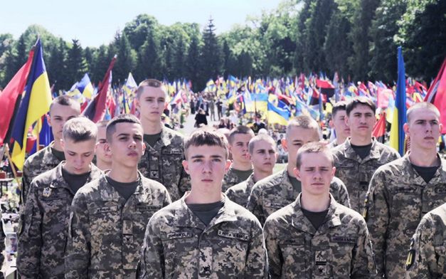 Soldiers stand in the sunlight. Ukrainian flags in the background. 