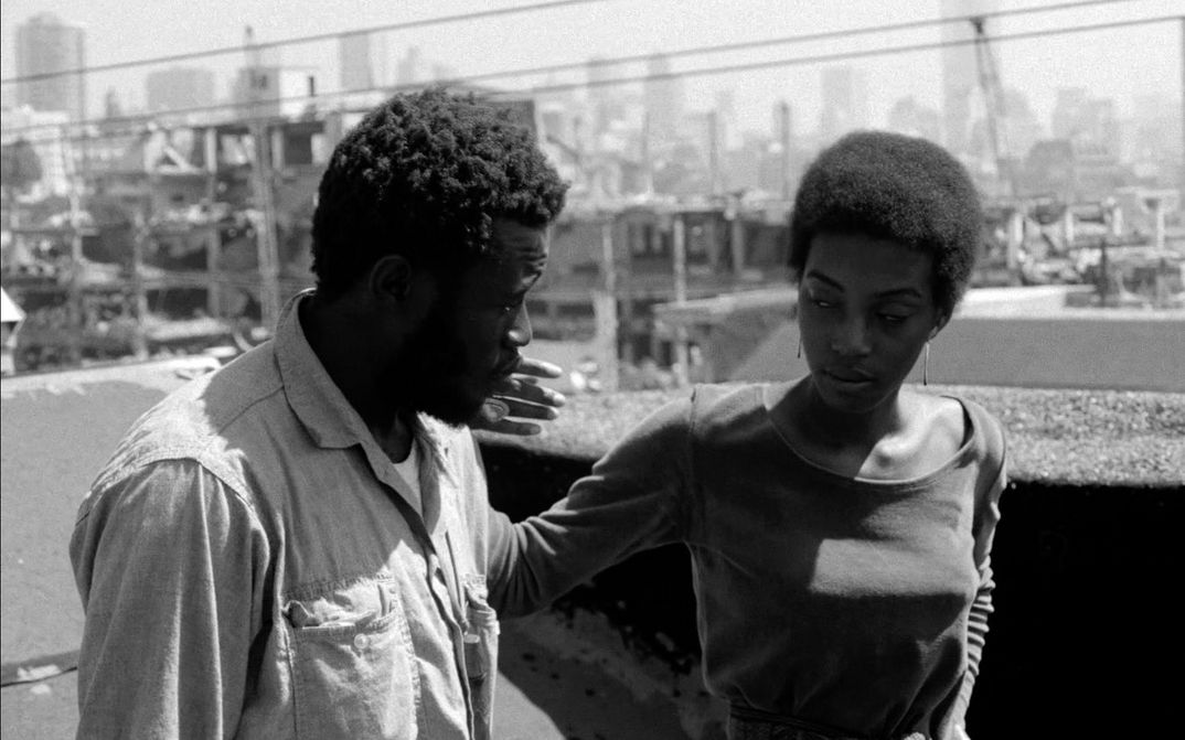 Film still from BUSHMAN: A man and a woman walk along a street, she strokes his cheek.