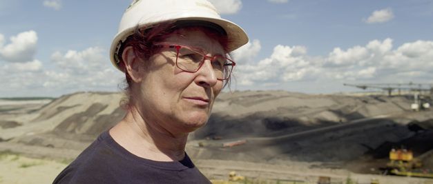 Filmstill aus STOLZ & EIGENSINN von Gerd Kroske. Das Bild zeigt eine Frau mit Schutzhelm und roter Brille vor einem Tagebau.