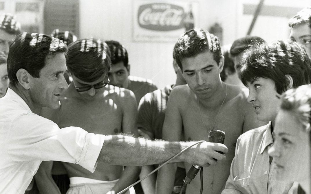 Filmstill aus COMIZI D´AMORE: Eine Gruppe von Menschen. Eine Mann hält ein Mikrofon zu einer Person.