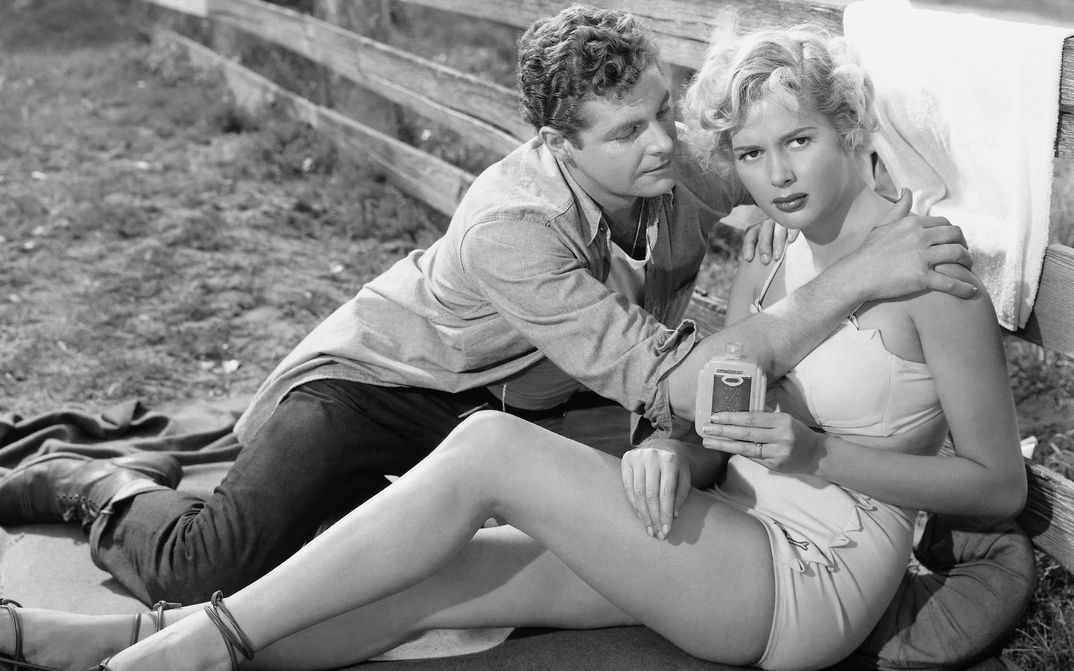Film still from PICKUCK: A young, scantily clad woman sits leaning against a fence. A man sits next to her and grabs her by the shoulders.