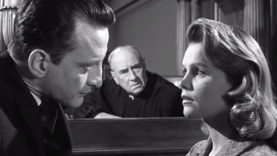 Filmstill from ANATOMY OF A MURDER: A man and a woman look at each other. The woman looks startled. Another man is sitting in the background looking at the two of them.