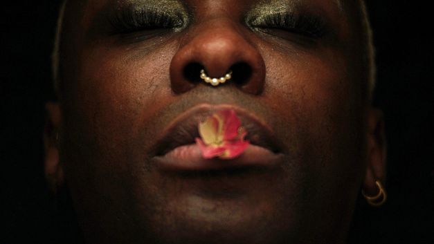 Extreme Nahaufnahme eines Gesichts mit geschlossenem Mund, Nasenpiercing und einer Blüte auf den Lippen.