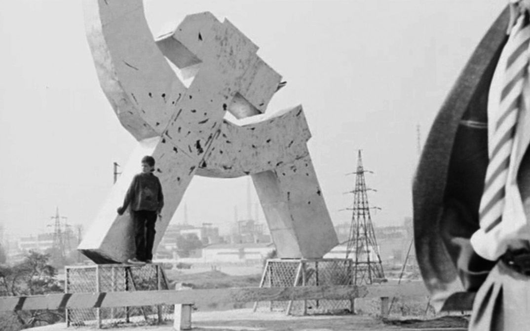 Film still from SWAN LAKE. THE ZONE: A person leans against a hammer and sickle monument.
