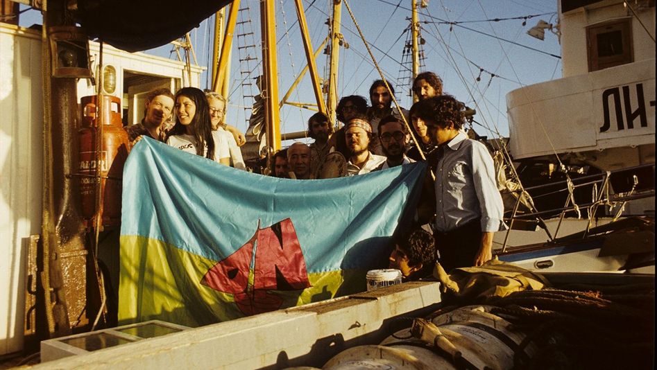 Filmstill aus OPERATION NAMIBIA: Eine Gruppe von Menschen steht auf einem Segelschiff. Sie sehen glücklich aus und halten eine Flagge in blau und gelb und einem roten Segelschiff in der Hand.