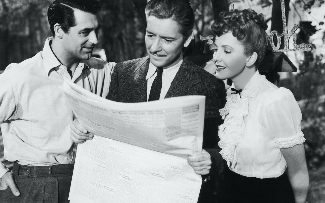 Film still from THE TALK OF THE TOWN: Two men and a woman stand together and look at a newspaper.
