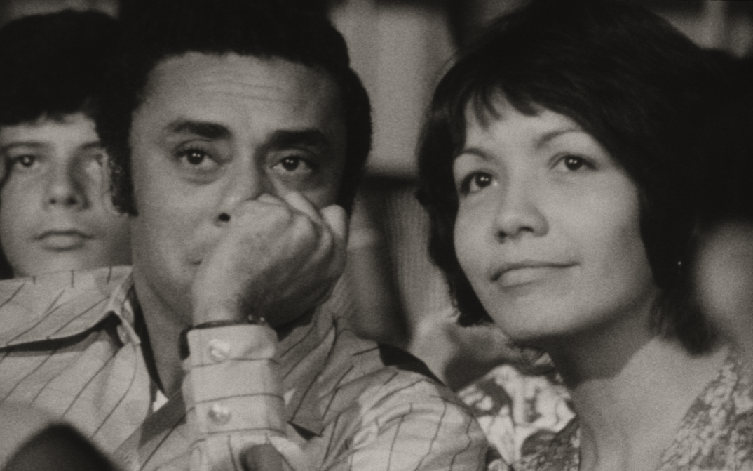 Film still from DE CIERTA MANERA: A woman and a man sit side by side and look at a screen off-screen.
