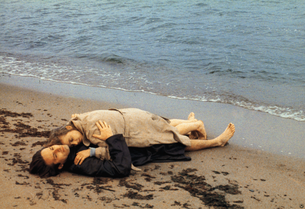 Film still from FLUCHT IN DEN NORDEN: A man and a woman are lying clothed on the beach. She lies on top of him and is embraced by him.