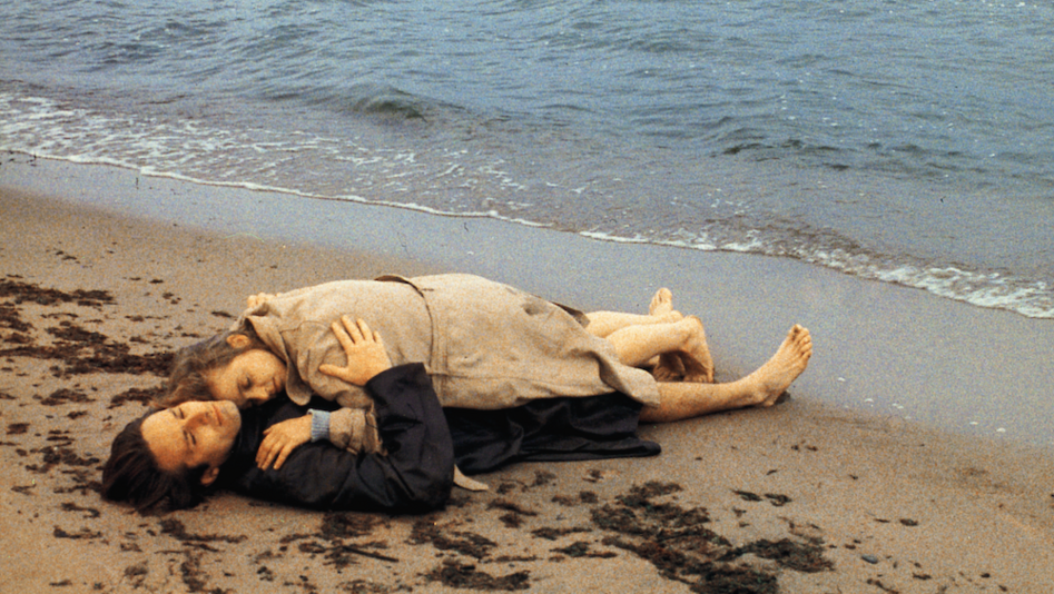 Film still from FLUCHT IN DEN NORDEN: A man and a woman are lying clothed on the beach. She lies on top of him and is embraced by him.