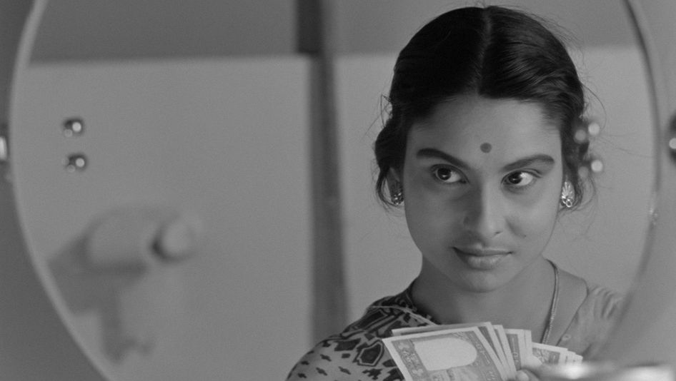 Filmstill from MAHANAGAR: A woman with a dot on her forehead looks in the mirror and holds a stack of banknotes in her hands.