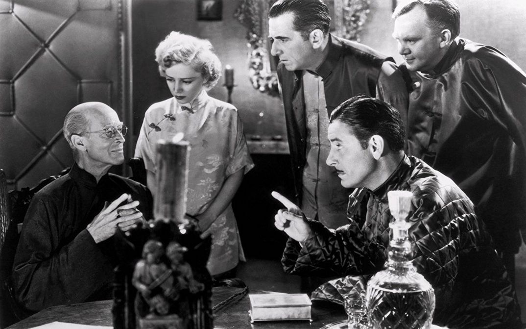 Film still from LOST HORIZON: A group of people are sitting and standing around a table. Everyone is looking at one person.