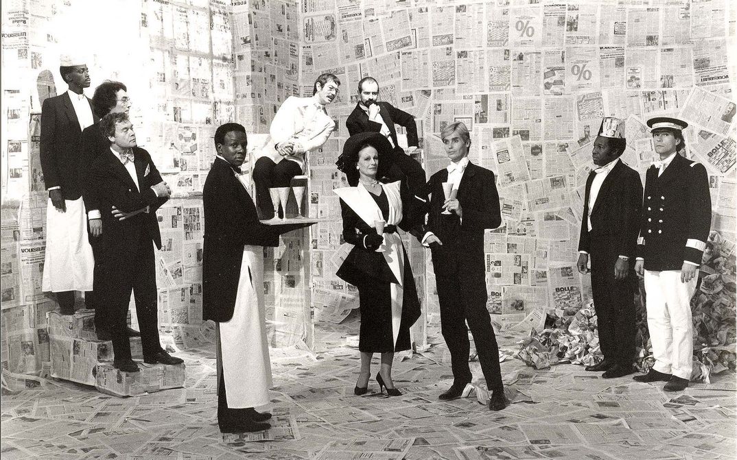 Film still from DORIAN GRAY IM SPIEGEL DER BOULEVARDPRESSE: A group of elegantly dressed people and a few waiters stand in a room completely lined with newspapers.