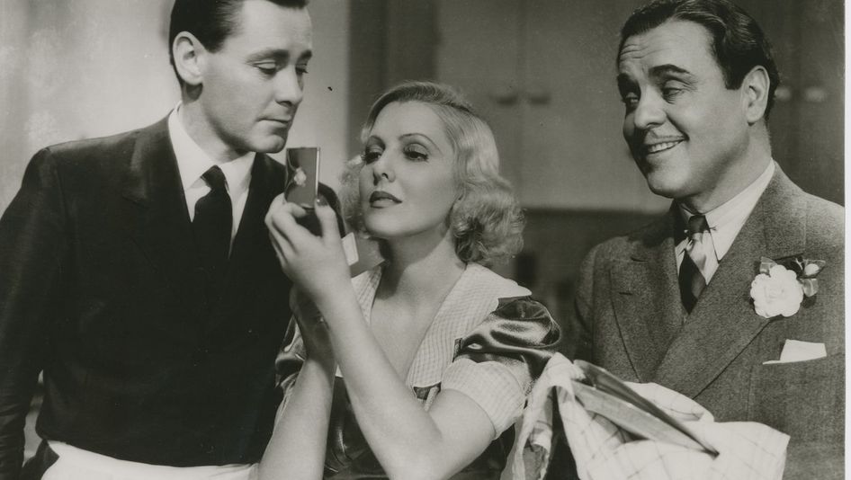 Film still from IF YOU COULD ONLY COOK: Two men and a woman are standing in a kitchen. The men are wearing aprons, one is drying dishes. The woman is looking in a hand mirror.