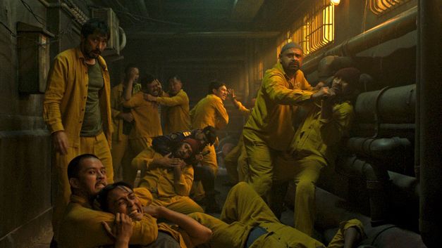 A group of men in yellow clothing fist fight with each other in a narrow, dark corridor lined with pipes.