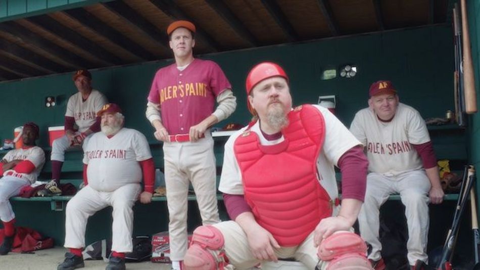 Filmstill aus EEPHUS: Eine Gruppe von Männern unterschiedlichen Alters, die alle Baseballkleidung tragen, schauen einem Spiel zu. 