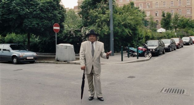 Filmstill from WANKOSTÄTTN: A man in a grey suit, tie and black top hat is leaning on an umbrella. He is standing in the street. In the background are parked cars, trees and houses.
