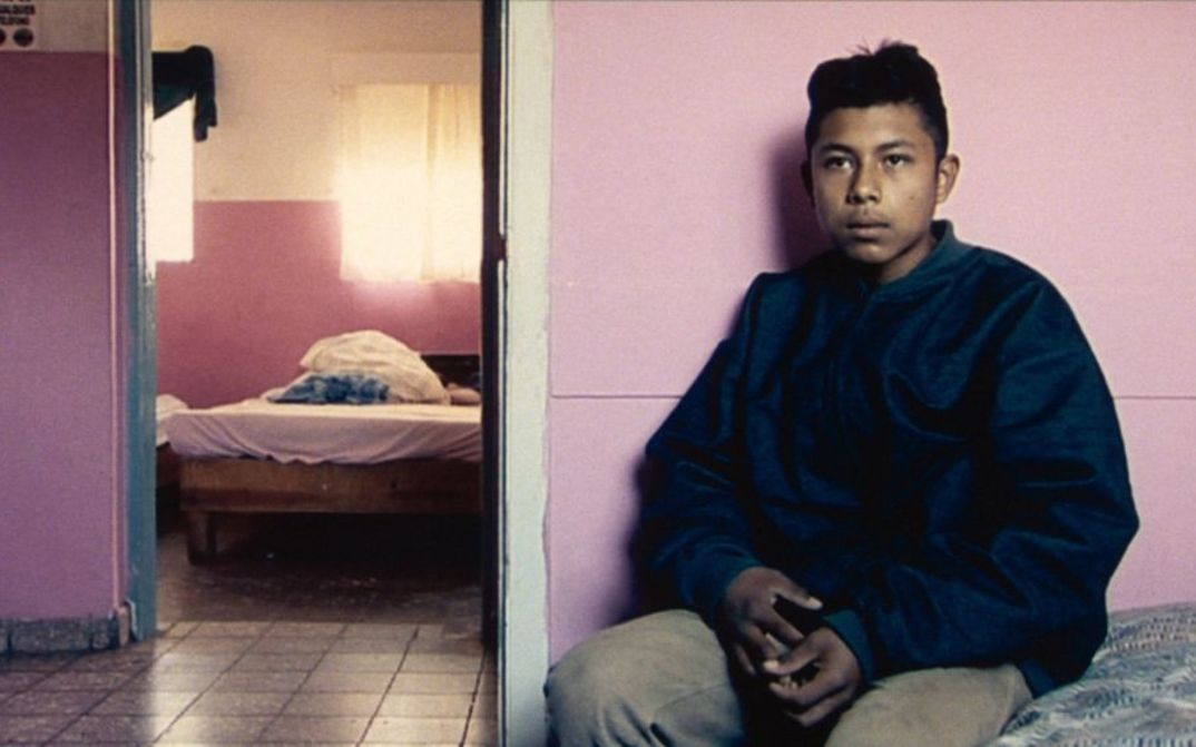 Film still from DE L’AUTRE CÔTÉ: A young man sits on a bed in sparse accommodation and looks into the camera.