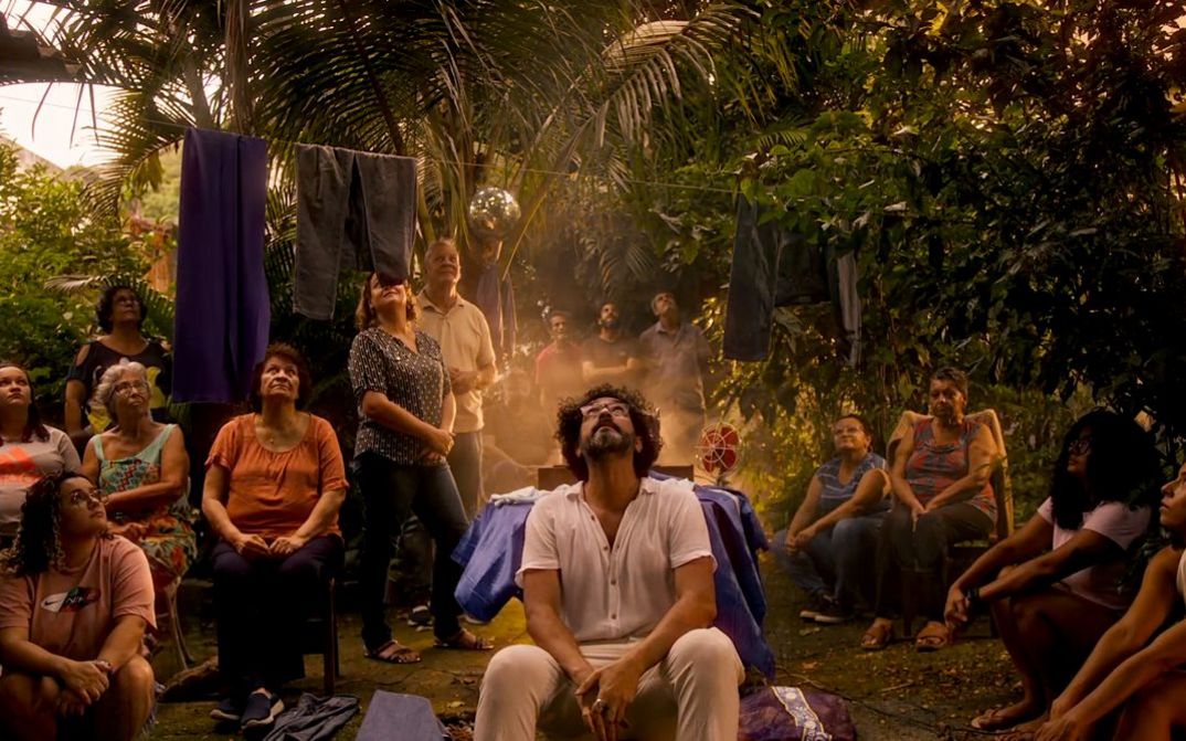 Film still from Felipe M. Bragança’s film “Zizi (ou oração da jaca fabulosa)”. In the courtyard of a house with abundant vegetation, an intergenerational group gazes at the sky. The protagonist sits at the center of the group while a golden, ethereal light rises from the ground, bathing the scene in a supernatural aura.