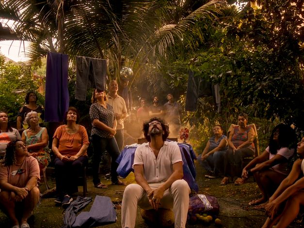 Film still from Felipe M. Bragança’s film “Zizi (ou oração da jaca fabulosa)”. In the courtyard of a house with abundant vegetation, an intergenerational group gazes at the sky. The protagonist sits at the center of the group while a golden, ethereal light rises from the ground, bathing the scene in a supernatural aura.