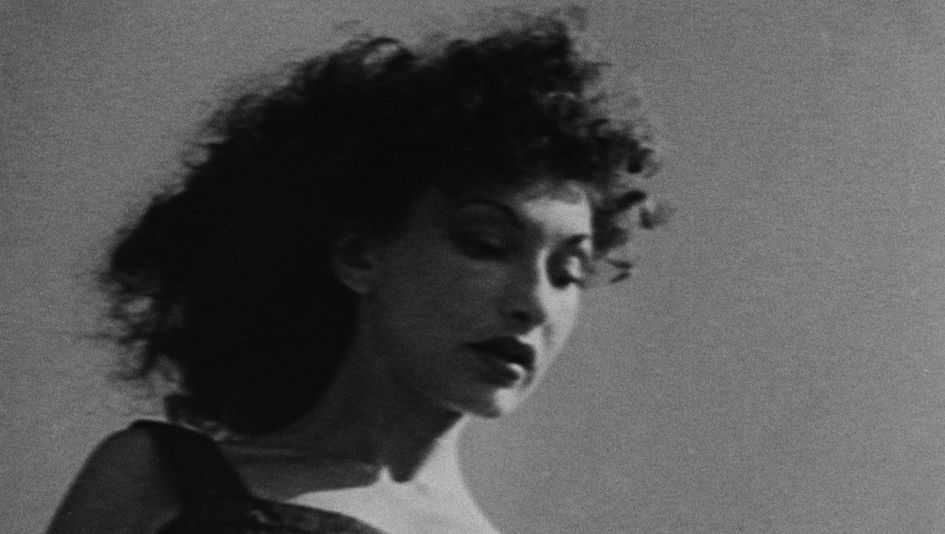 Filmstill aus AT LAND: Schwarz-weiß-Aufnahme einer Frau mit dunklen Locken, in leichter Untersich aufgenommen.