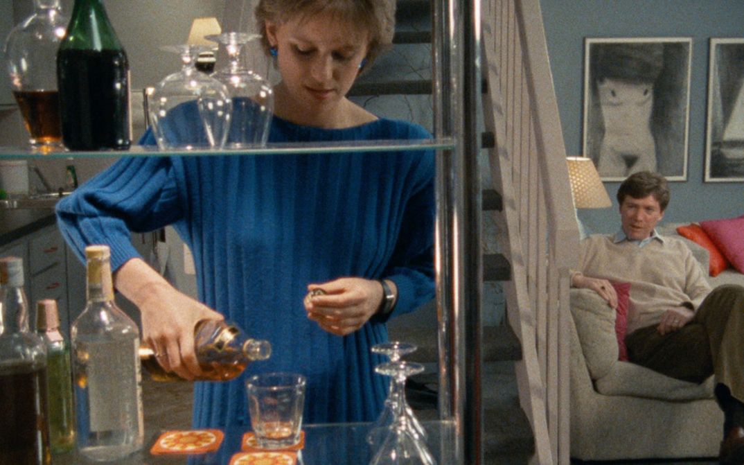 Film still from WORKING GIRLS. An elegantly dressed woman pours a drink in an apartment, in the background a man sits on a sofa.