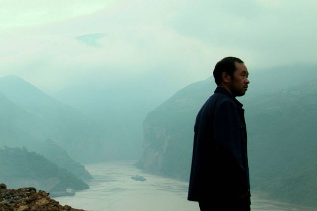 Filmstill from STILL LIFE: A man stands on high ground and looks to the right. A river and mountains below him. It is foggy.