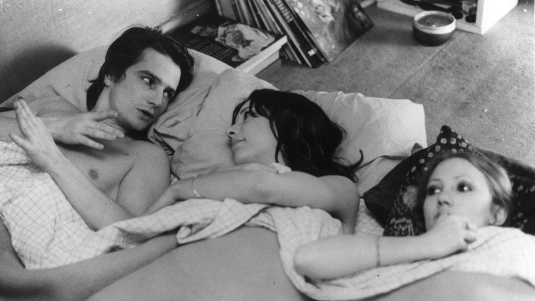 Filmstill aus LA MAMAN ET LA PUTAIN: Ein Mann und zwei Frauen liegen nebeneinander in einem Bett und unterhalten sich.