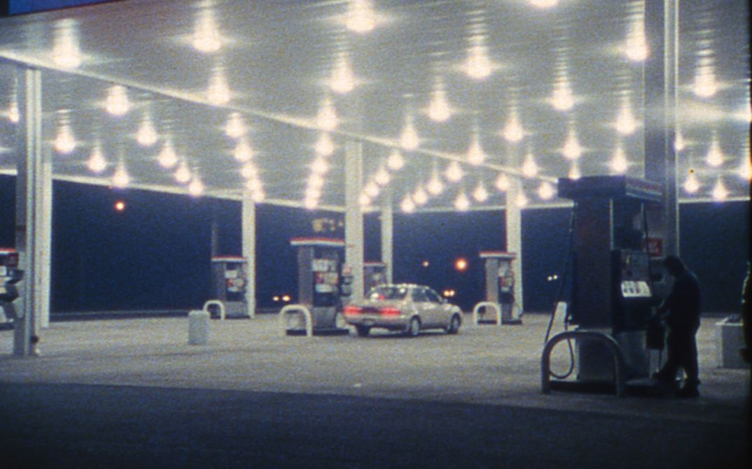 Filmstill from SOMETHING MORE THEN NIGHT: A petrol station in the dark. A car is parked at the petrol pump in the middle.