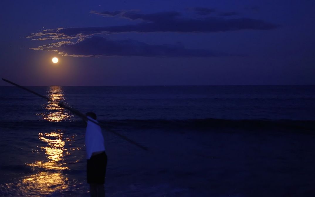 Filmstill aus TIME FLOWS ALL OVER: Man sieht das vom Mond beschienene Meer. Im Vordergrund steht eine Person.