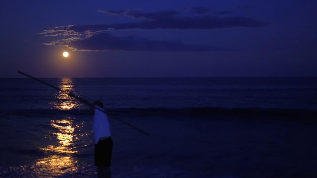 Film still from TIME FLOWS ALL OVER: You can see the sea illuminated by the moon. A person stands in the foreground.