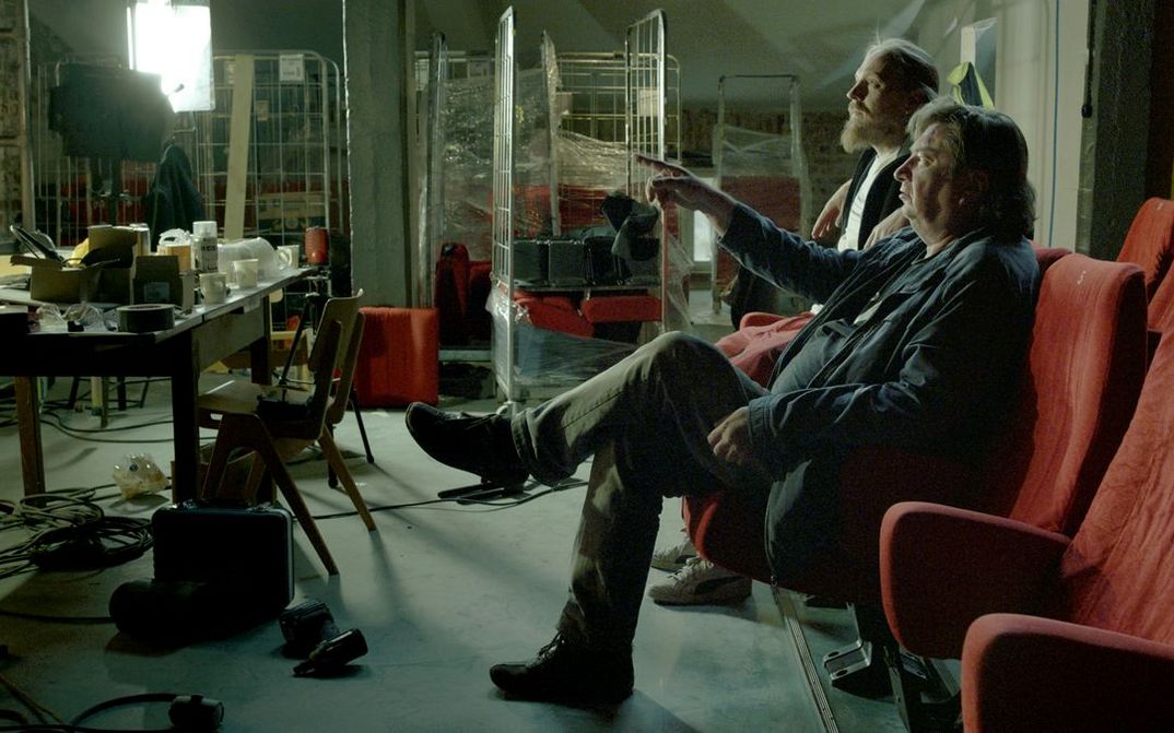 Film still from CINÉMA LAIKA: Two men are sitting on movie theater chairs in a somewhat chaotically cluttered room. One of them is the director Aki Kaurismäki.