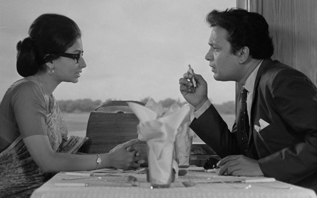 Filmstill from NAYAK: A man and a woman are sitting at a restaurant table by the window. The man seems impulsive, he is holding a cigarette in his hand.