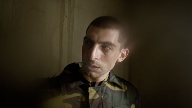 Film still from AFTER DREAMING by Christine Christine Haroutounian. Close-up of a soldier in uniform in front of a wall. 