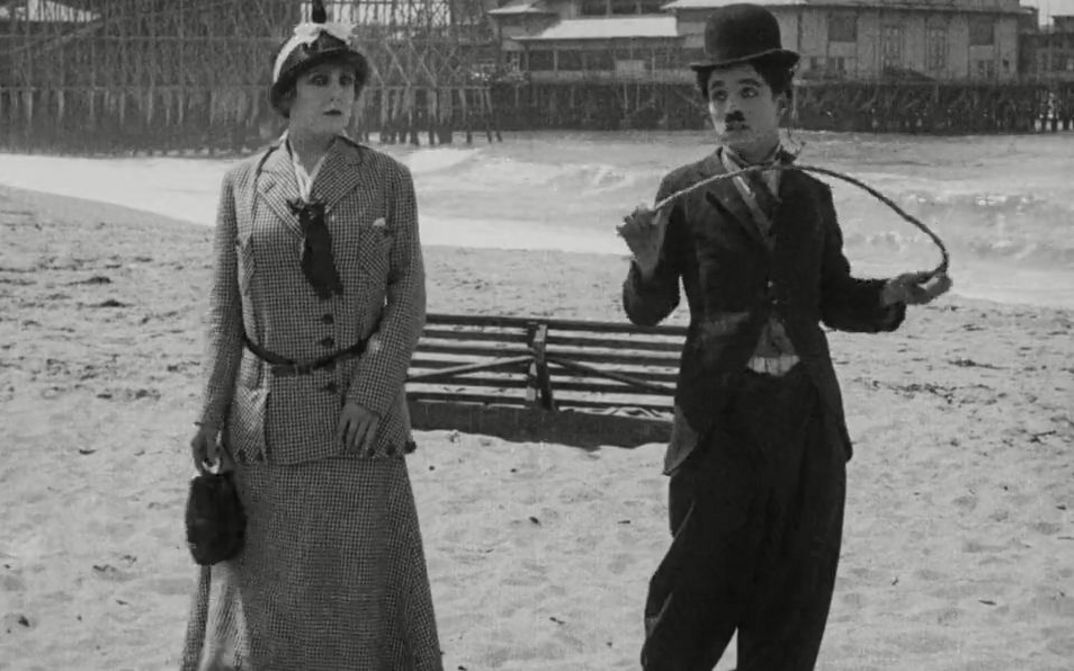 Film still from BY THE SEA: Charlie Chaplin and a woman standing next to each other on the beach.