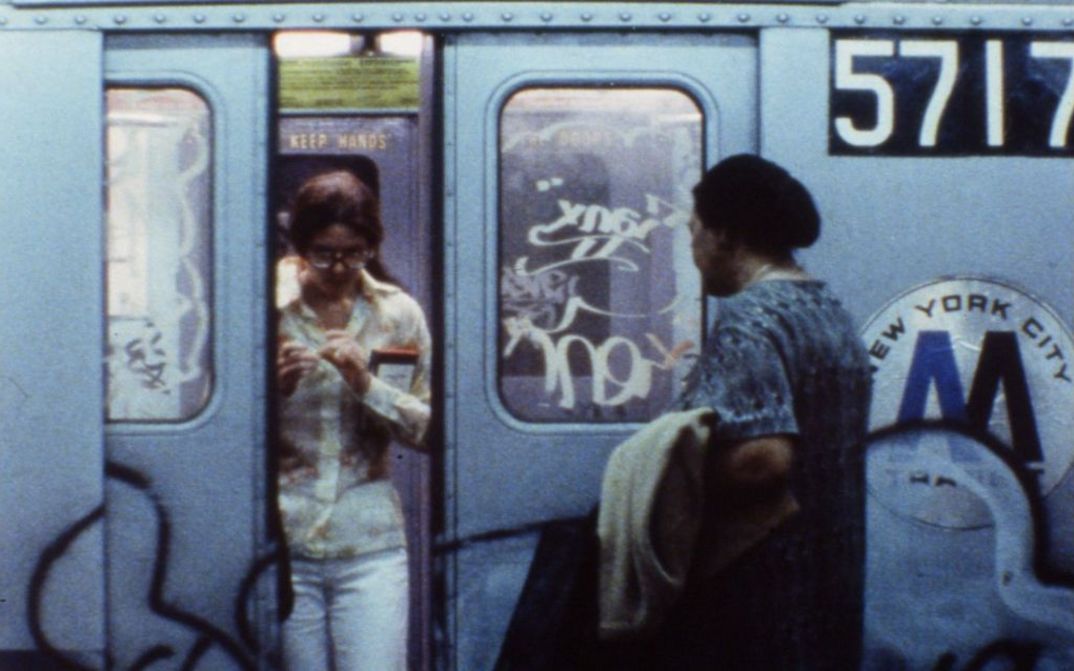 Film still from NEWS FROM HOME: he graffiti-covered doors of a New York subway open, one woman gets off, another gets on.