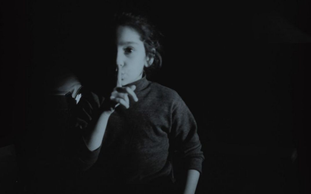 Film still from MES FANTÔMES ARMENIENS by Tamara Stepanyan. The black and white image shows a girl placing the index finger of her right hand in front of her lips.
